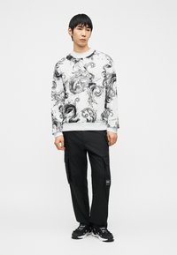 Άνδρας που στέκεται φορώντας λευκό φούτερ με μαύρα κυματιστά σχέδια, μαύρο παντελόνι καμπαρντίνα και μαύρα αθλητικά παπούτσια με χοντρές λευκές σόλες.