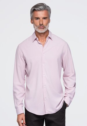 Homme aux cheveux gris et à la barbe, portant une chemise rose clair à manches longues boutonnée et un pantalon noir, debout devant un fond uni.