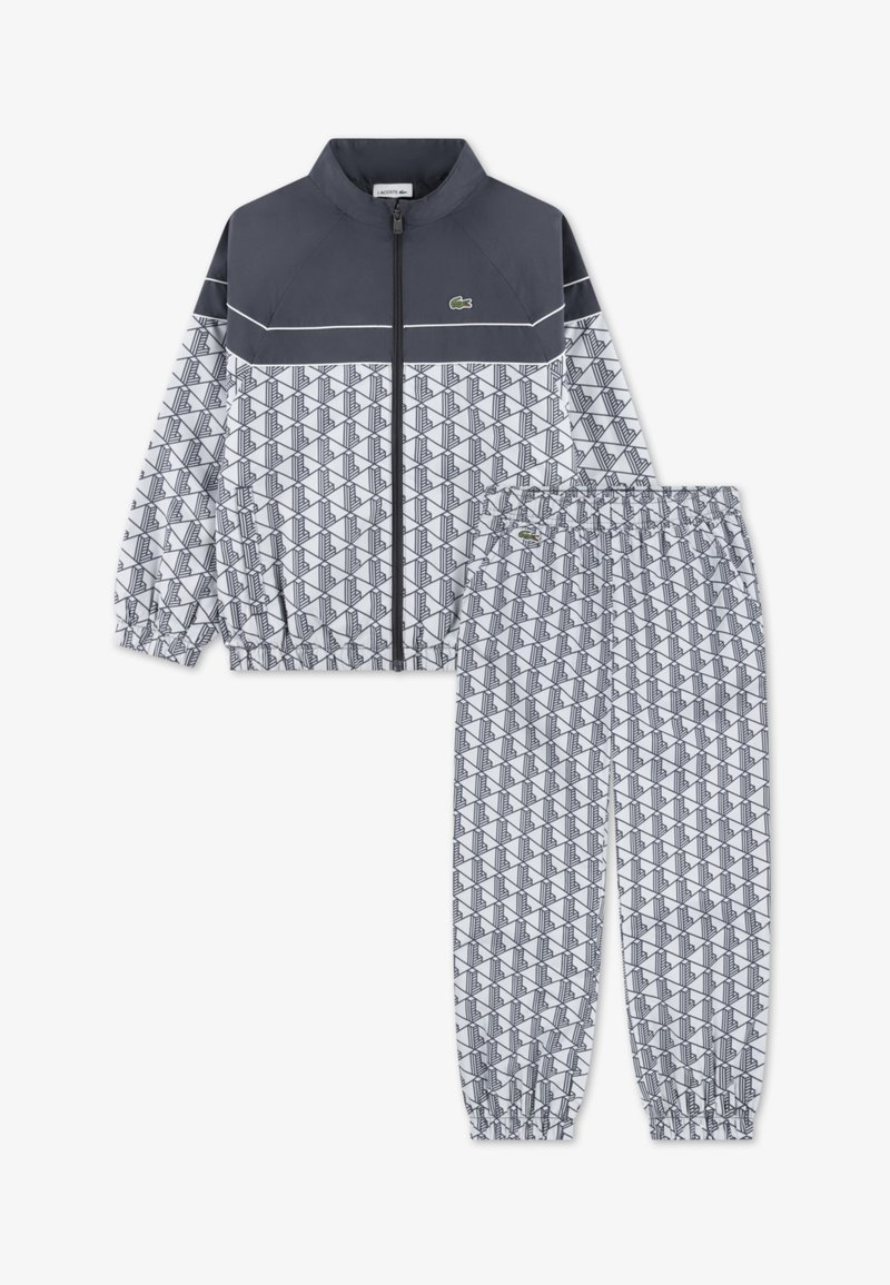 Trenerka z zgornjim delom na zadrgo in hlačami z elastičnim pasom. Temno sivi zgornji del, beli spodnji del z geometrijskim vzorcem, z logotipom Lacoste.