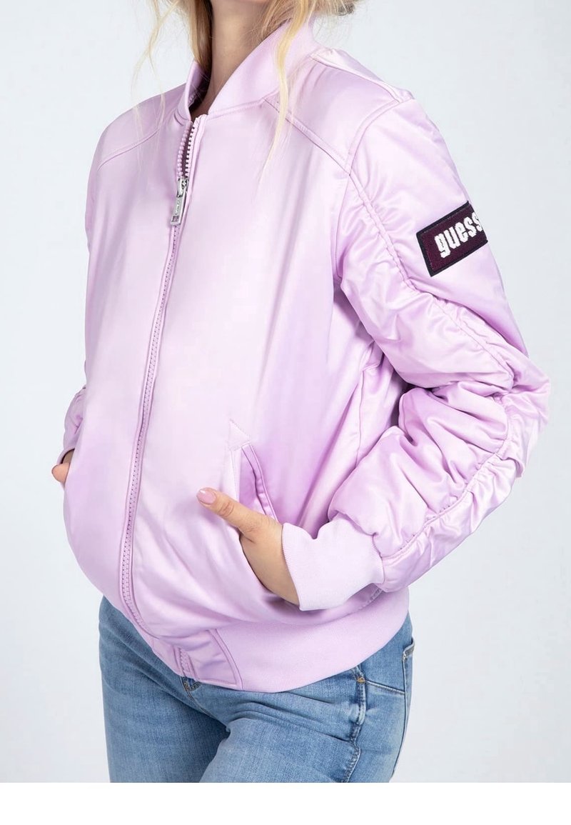 Giubbotto bomber lilla con chiusura a zip, polsini a coste e due tasche laterali. Presenta un patch con logo sul braccio sinistro e una texture liscia e lucida.