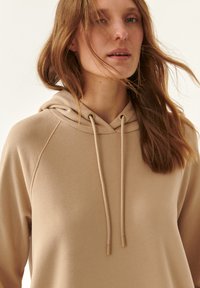 Sudadera beige con capucha y cordones, con mangas raglán y una tela suave y texturizada. Detalles sutiles alrededor del escote y los puños.