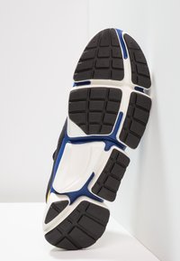 Černo-bílá podešev tenisky s modrými a žlutými akcenty. Má texturovaný vzor pro záběr a zakřivený design pro flexibilitu.