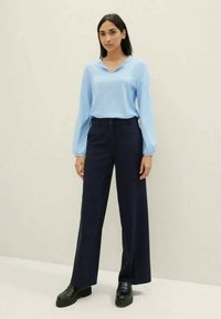 Woman standing wearing a light blue long-sleeve blouse, navy wide-leg trousers, and black platform shoes against a plain background.