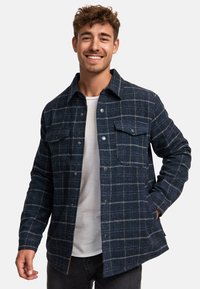 Blue plaid jacket made of textured fabric, featuring a collared neck, front pockets, and button closure. Worn over a white shirt.
