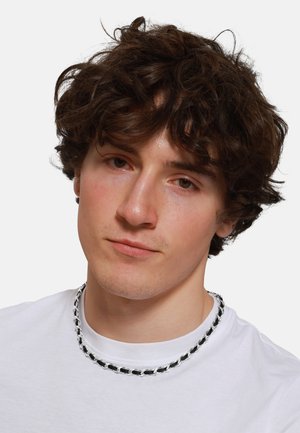 Silver and black chain necklace with interlocking links, worn on a plain white t-shirt; soft curls visible around the neckline.