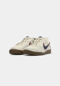 Białe buty sportowe o niskim profilu, z chropowatą skórzaną cholewką, ciemnoniebieskim logo Nike oraz brązową gumową podeszwą.