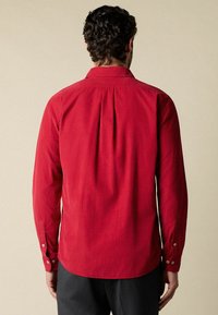 Man met krullend haar, gekleed in een rode lange mouwen shirt, gezien vanaf de achterkant, staand tegen een effen beige achtergrond.