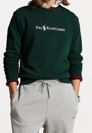 Personne portant un sweat-shirt Polo Ralph Lauren vert foncé et un pantalon de survêtement gris clair, les mains dans les poches et une montre au poignet gauche.