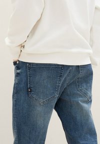 Jean en denim bleu à coupe droite, avec deux poches arrière et des coutures d'accent. Le tissu a une apparence usée et texturée.