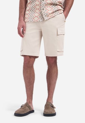 Man draagt beige cargoshorts, een shirt met korte mouwen met patroon en bruine suède instapschoenen, staat met één hand in de zak.