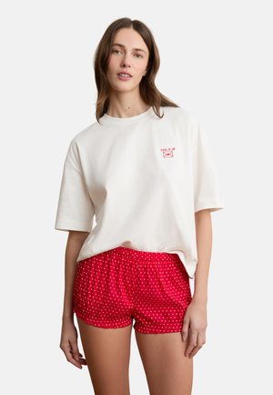 Junge Frau trägt ein weißes, übergroßes T-Shirt mit rotem Text und roten Shorts mit Punkten, steht vor einem schlichten weißen Hintergrund.