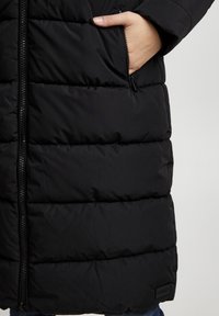 Doudoune noire avec un design matelassé, fermeture éclair à l'avant et poche latérale. Le matériau a une texture lisse avec un accent de marque près de l'ourlet.