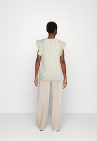 Blusa texturada cinza claro com mangas em babados, combinada com calças largas em bege. O conjunto apresenta um corte descontraído e detalhes discretos salpicados.