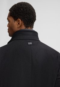 Uomo con capelli ricci corti che indossa un cappotto nero a collo alto, di profilo mostrando la schiena e un'etichetta metallica al collo.