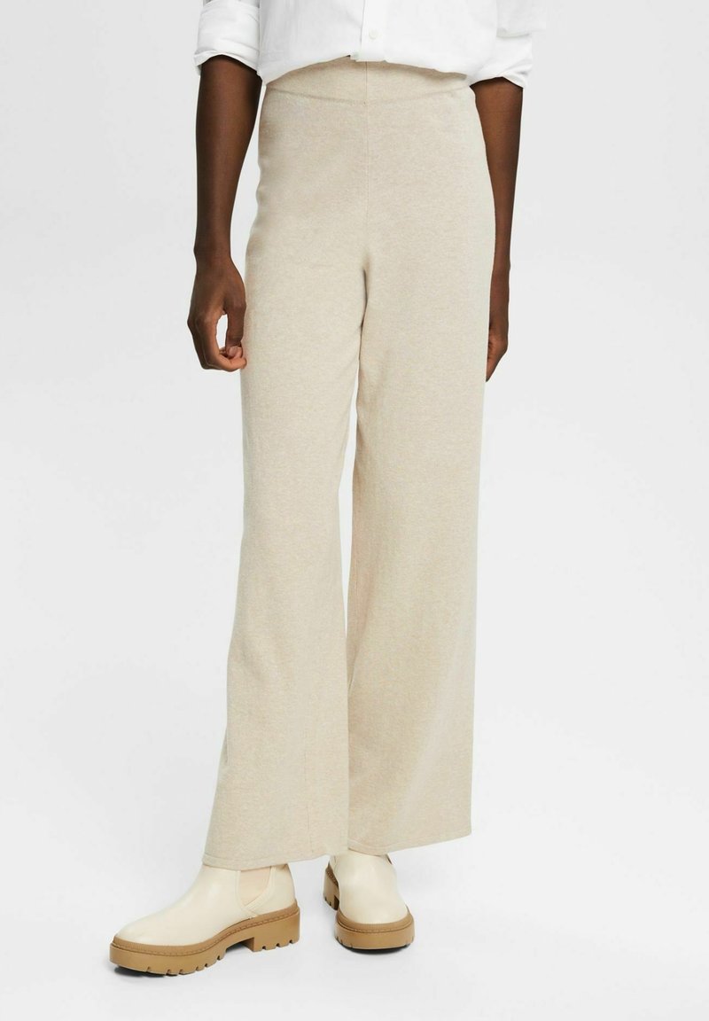 Personne portant un pantalon beige à jambes larges, une chemise blanche aux manches retroussées et des bottines crème à semelle épaisse sur fond blanc.