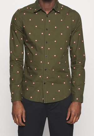 Mężczyzna w oliwkowo-zielonej koszuli na guziki z długimi rękawami i małymi różowymi kropeczkami, w połączeniu z czarnymi spodniami.