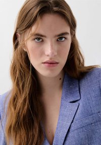 Blauwe blazer van een gestructureerde stof met subtiele, glanzende deeltjes. Opvallende notch-lapel en een enkele borstzak. Model staat recht voor de camera.