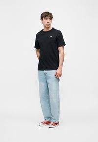 Νέος άνδρας που στέκεται φορώντας μαύρο μπλουζάκι, ανοιχτόχρωμο μπλε τζιν και κόκκινα αθλητικά παπούτσια μπροστά σε λευκό, απλό φόντο.