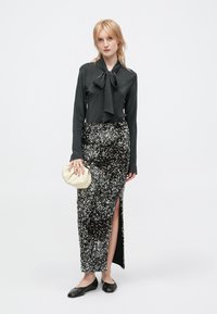 Donna che indossa una blusa nera con un fiocco, una gonna di paillettes argento con uno spacco sul coscia, tacchi bassi neri e tiene in mano una piccola pochette beige.