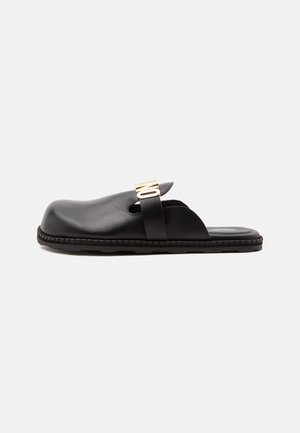 Chaussure slip-on en cuir noir avec une semelle épaisse et une large bande ornée de lettres "NO" dorées sur le dessus.