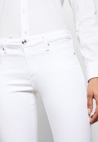 Jeans bianchi in denim con una vestibilità slim, design a cinque tasche, accessori in metallo argentato e dettagli di cucitura a contrasto. Abbinati a una camicia bianca con bottoni.