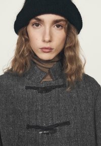 Cappotto di lana grigio con colletto alto, chiusure a fibbia e tessuto strutturato. Indossato sopra un dolcevita marrone e abbinato a un cappello nero.