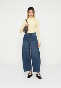 Suéter de cuello alto amarillo de manga larga, jeans de denim de tiro alto y pierna ancha, zapatos negros de tacón cuadrado y un pequeño bolso oscuro.