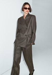 Blazer marrone doppiopetto con bottoni dorati, abbinato a pantaloni coordinati. Vestibilità slim e su misura con texture liscia. Occhiali da sole scuri inclusi.