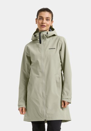 Kobieta w jasnozielonym wodoodpornym płaszczu Didriksons z kapturem i przednim zamkiem, stojąca na jednolitym tle.