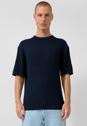 Mężczyzna z krótkimi włosami ubrany w granatowy prążkowany sweter z krótkim rękawem i jasnoniebieskie jeansy, stojący na tle jednolitego szarego tła.