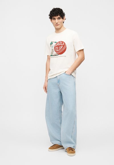 Νέος άνδρας με σγουρά σκούρα μαλλιά, που φοράει λευκό t-shirt με σχέδιο, ανοιχτό μπλε παντελόνι με φαρδιά μπατζάκια και καφέ αθλητικά παπούτσια, μπροστά σε αφηρημένο φόντο.