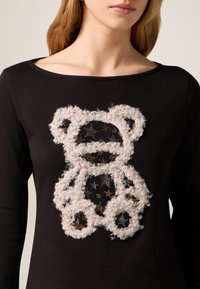 Czarna koszula z długim rękawem z wzorem misia zrobionego z puszystego, białego materiału z metalicznymi złotymi gwiazdkami w tle.