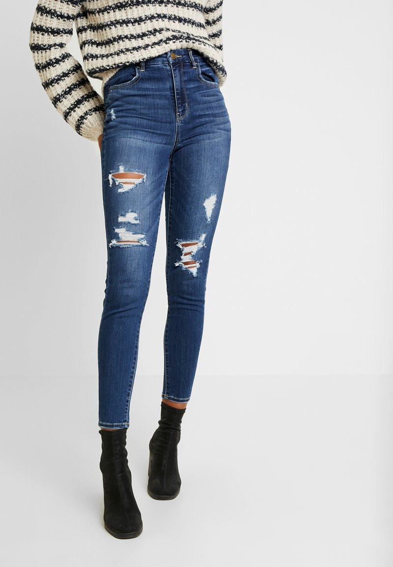 Smal passform blå rivna jeans som bärs med en kräm-och-svart randig tröja och svarta ankelboots mot en vit bakgrund.