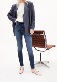 Harmaa bleiseri valkoisen neuleen päällä, yhdistetty kapeisiin sinisiin farkkuihin ja vaaleanpunaisiin kenkiin. Lähellä on moderni krominen tuoli ruskealla pehmustetulla istuimella.