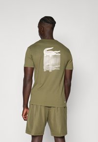 Ελιά πράσινη βαμβακερή μπλούζα T-shirt με ένα λευκό γραφικό κροκοδείλου και σχέδιο κατακόρυφων γραμμών στην πλάτη. Βραχίονες κοντοί, στρογγυλή λαιμόκοψη και άνετη εφαρμογή.