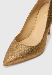 Zlatna sjajna štikla s šiljastim vrhom, glatkim unutrašnjim dijelom i teksturiranim vanjskim dijelom. Ima stiletto potpeticu i moderan dizajn.