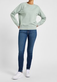 Sudadera verde claro con cuello redondo y puños acanalados, combinada con vaqueros ajustados azul oscuro y zapatillas blancas. La tela parece suave con una textura lisa.