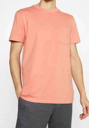 Mies, joka on pukeutunut lohenväriseen lyhythihaiseen t-paitaan, jossa on rintatasku, ja tummiin teksturoiduihin housuihin valkoista taustaa vastaan.