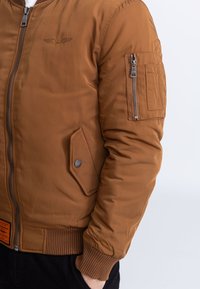 Bruine bomberjas met een ritssluiting aan de voorkant, geribbelde manchetten en een zakje op de mouw. Heeft een logo en een zijzak met drukknopsluiting.