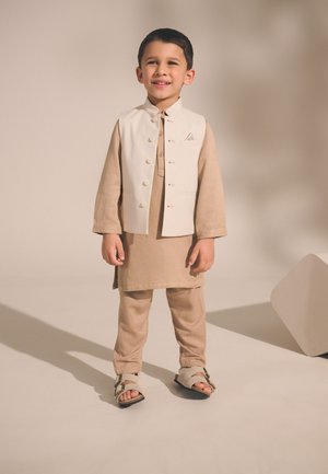 Beżowa kurta i spodnie z białą kurtką z guzikami, w połączeniu z beżowymi sandałami. Gładka faktura tkaniny i prosty design.