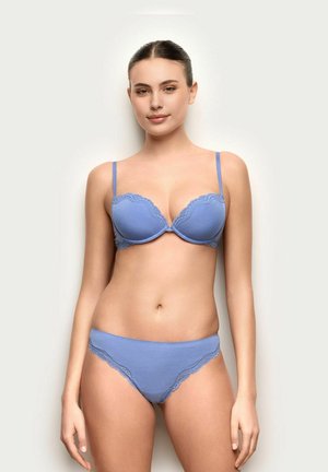 Mujer de cabello oscuro con un sujetador azul claro con encaje y ropa interior a juego, de pie frente a un fondo claro y liso.