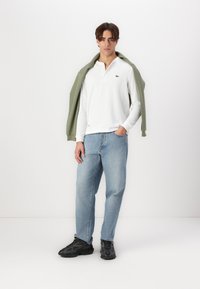 Weiße Langarm-Poloshirt mit Kragen und grünen Akzenten, kombiniert mit hellblauen Jeans und schwarzen Sneakers, abgerundet mit einem grünen Pullover.