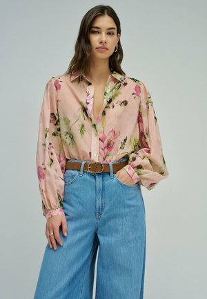 Femme portant un chemisier transparent rose à fleurs, rentré dans un jean bleu taille haute à jambes larges, avec une ceinture marron, posant devant un fond uni.