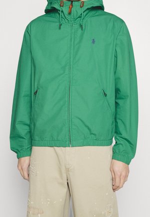 Mann trägt eine grüne Kapuzenjacke mit frontalem Reißverschluss und Taschen, kombiniert mit beige-zerfetzten Hosen.