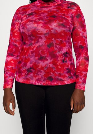 Personne portant un haut à manches longues et col montant avec un motif floral rouge et rose et un pantalon noir, montré des épaules aux hanches.