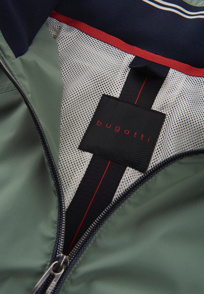 Veste légère verte avec un intérieur en mesh, dotée d'une étiquette noire avec du texte rouge, et d'un col marine avec des rayures d'accent rouges.