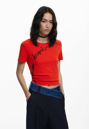T-shirt rouge en coton à manches courtes avec une coupe ajustée, featuring un texte et un design noir. Assorti à un pantalon foncé et une ceinture bleu contrastée.