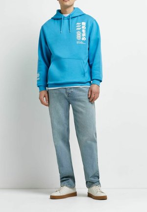 Uomo con felpa con cappuccio di un azzurro brillante con scritte grafiche bianche, jeans azzurri chiari e scarpe da ginnastica bianche, in piedi su sfondo bianco uniforme.