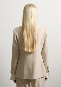 Persoon met lang blond haar, gekleed in een beige colbert en bijpassende broek, staande tegen een effen witte muur, met de rug naar de kijker.