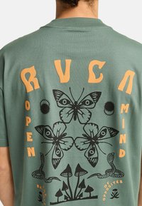 Camiseta de algodón verde con ilustraciones de mariposas negras y texto naranja que dice "MENTE ABIERTA", con elementos decorativos como serpientes y champiñones.